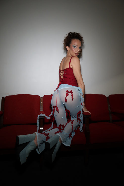 A woman reclining on a red theater-style seat, wearing a red corset top and a sheer blue pants with red bow, against a plain light gray wall.