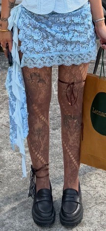 A person wearing a light blue floral lace mini skirt, sheer brown lace tights, and black platform loafers