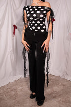 A model wearing a black and white polka dot off-the-shoulder top with sheer ruffled tie details on the sleeves and sides, paired with black wide-leg pants, standing against a white draped fabric backdrop.
