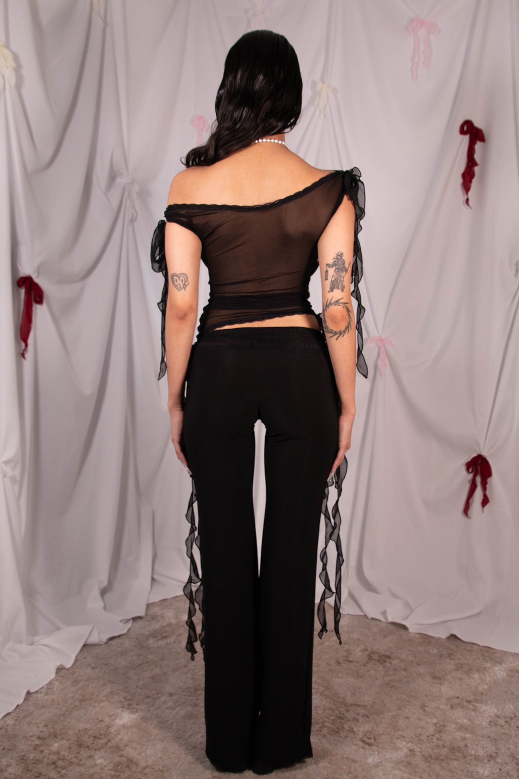 A woman shown from the back wearing a black sheer off-the-shoulder top with long ribbon ties and matching black wide-leg pants, standing in front of a draped white fabric backdrop with small red and pink ribbon decorations.