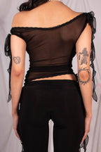 A woman shown from the back wearing a black sheer off-the-shoulder top with long ribbon ties and matching black wide-leg pants, standing in front of a draped white fabric backdrop with small red and pink ribbon decorations.