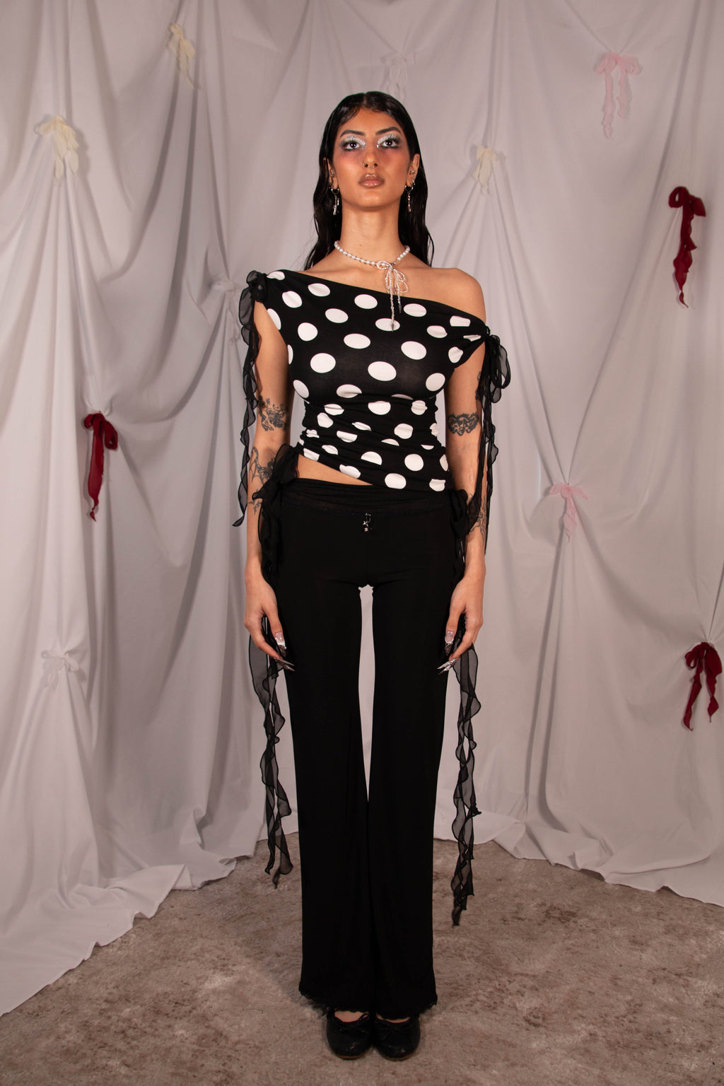 A model stands in front of a draped white fabric backdrop wearing a black off-the-shoulder crop top with white polka dots and sheer black ruffled straps, paired with black wide-leg pants.
