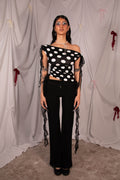 A model stands in front of a draped white fabric backdrop wearing a black off-the-shoulder crop top with white polka dots and sheer black ruffled straps, paired with black wide-leg pants.
