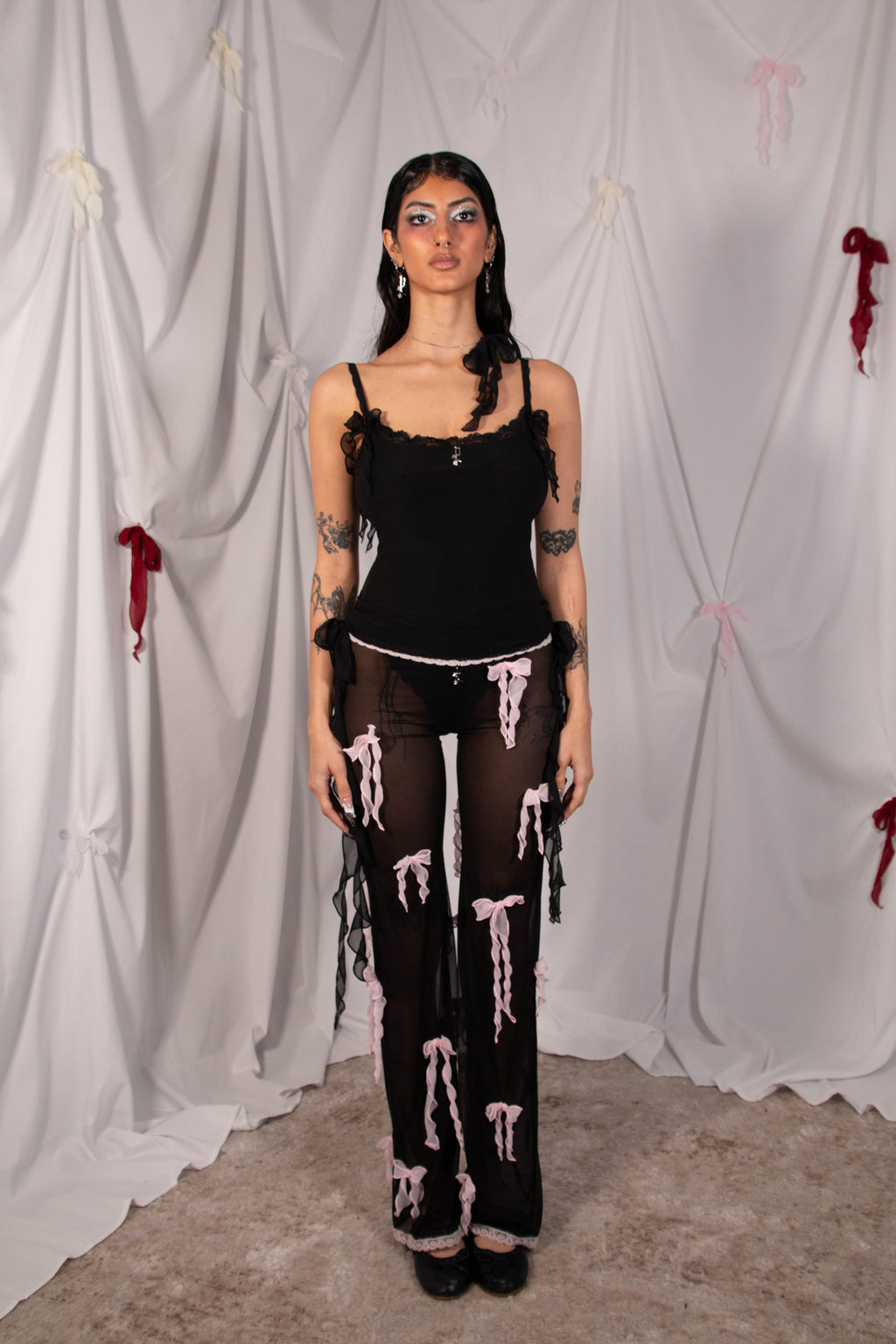A model stands against a draped white fabric backdrop wearing a black gothic-style outfit consisting of a black fringed camisole, a black sheer top with white dripping print, and matching black sheer pants with white dripping print.