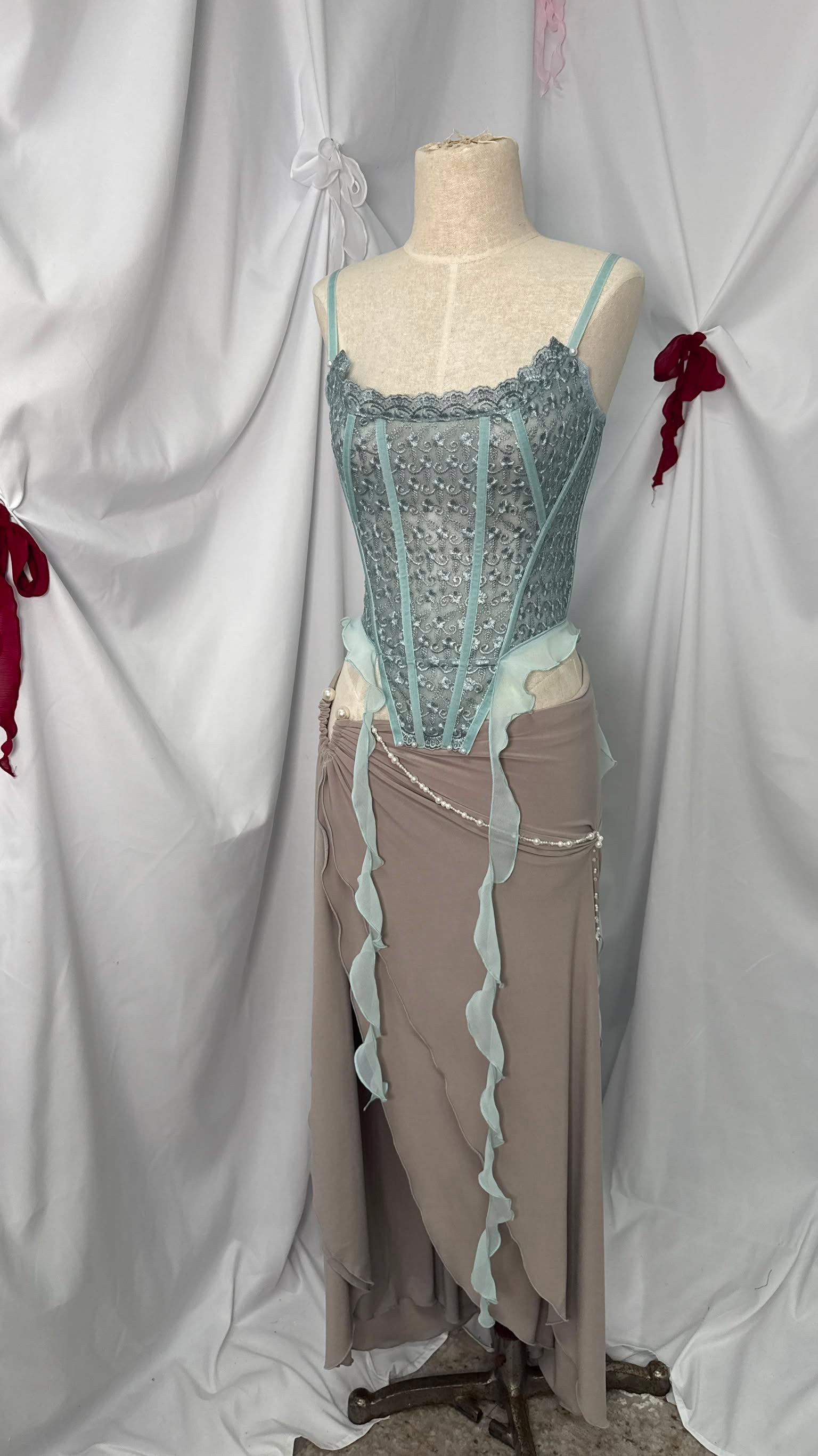 A costume-style outfit displayed on a dress form mannequin against draped white fabric. The top is a fitted lace corset in pale aqua with sheer trim, paired with a taupe skirt featuring pale aqua ruffled trim and a small waist chain.