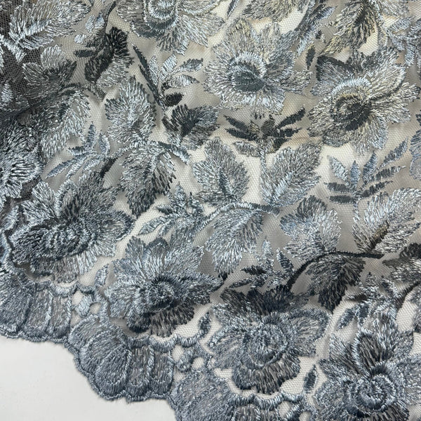 A close-up of a sheer floral lace fabric with embroidered rose motifs in gray, silver, and black tones over a light beige mesh backing.
