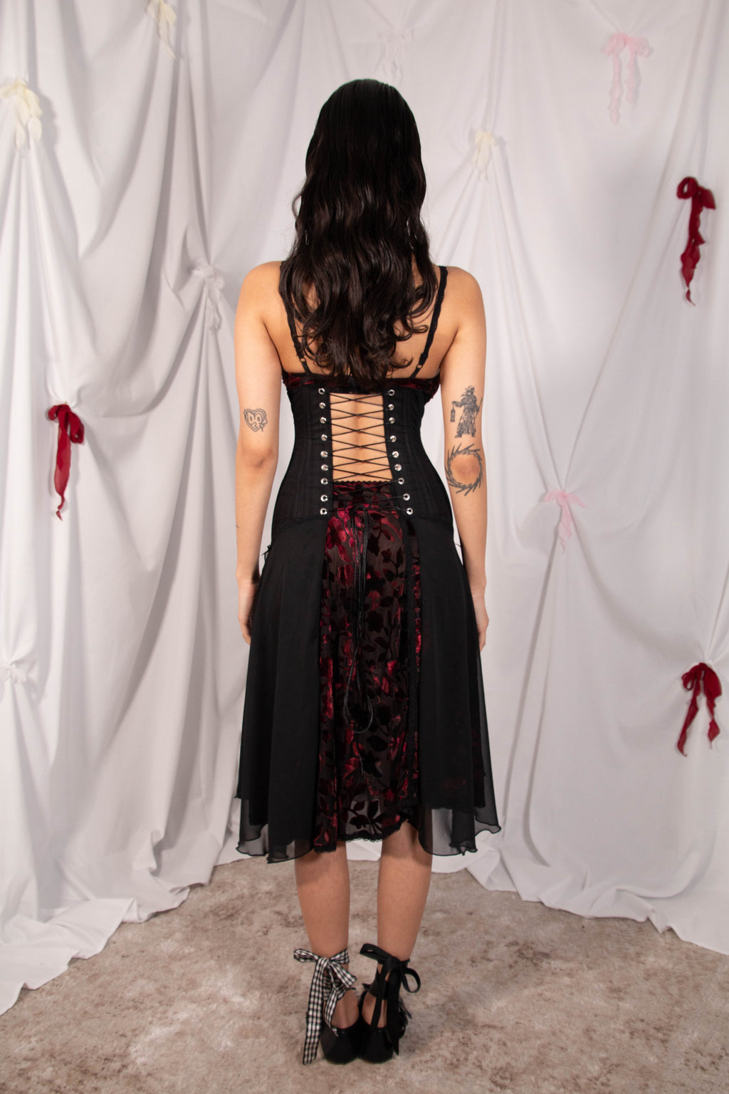 A woman stands with her back to the camera wearing a black gothic-style dress with a lace-up corset back, sheer black overlay, and a red floral tulle skirt. She is posed in front of a white draped backdrop with red ribbon accents.