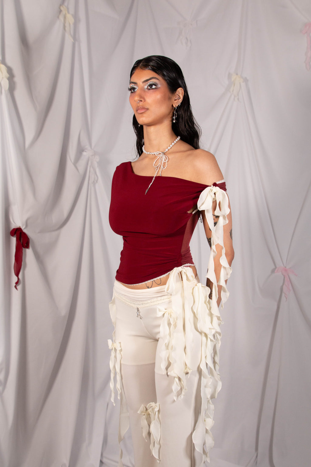 A model wearing a burgundy off-the-shoulder top with white tattered sleeve accents and white pants, standing against a draped white fabric backdrop.
