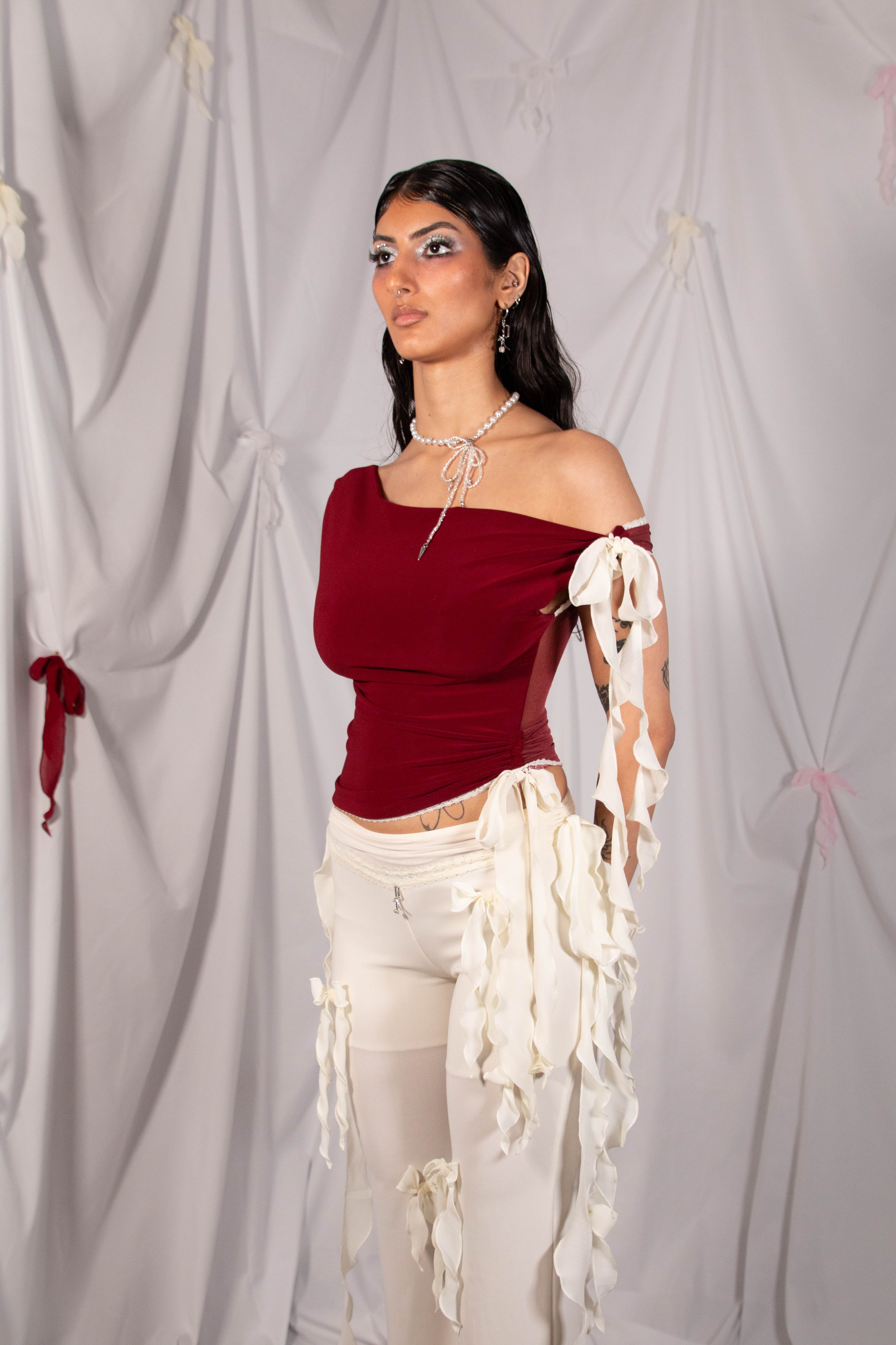 A model wearing a burgundy off-the-shoulder top with white tattered sleeve accents and white pants, standing against a draped white fabric backdrop.