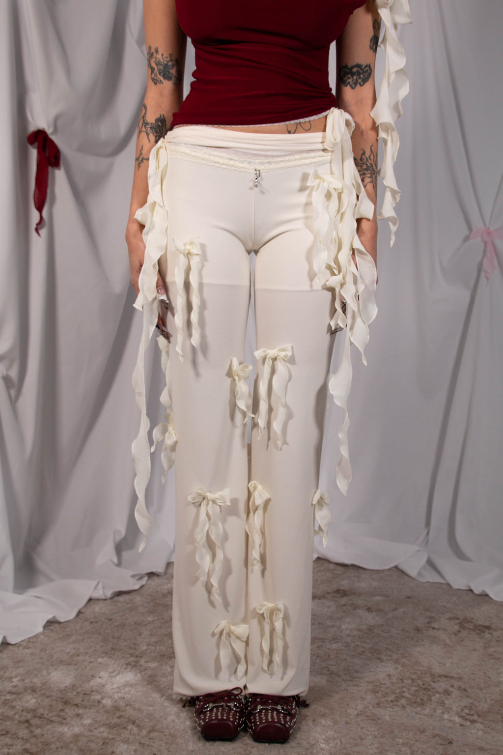 A model wearing off-white wide-leg pants with long draped ruffle trim and tie details, paired with a dark red top and brown lace-up shoes, standing against a draped white fabric backdrop.