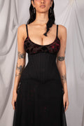 A woman modeling a black corset-style dress with a sheer dark floral lace bust and a flowing black skirt, posed against a draped light gray fabric backdrop.