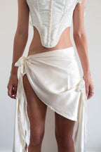 A model wearing a cream-colored satin wrap skirt tied at the hips with long draped tails, paired with a matching lace corset top.