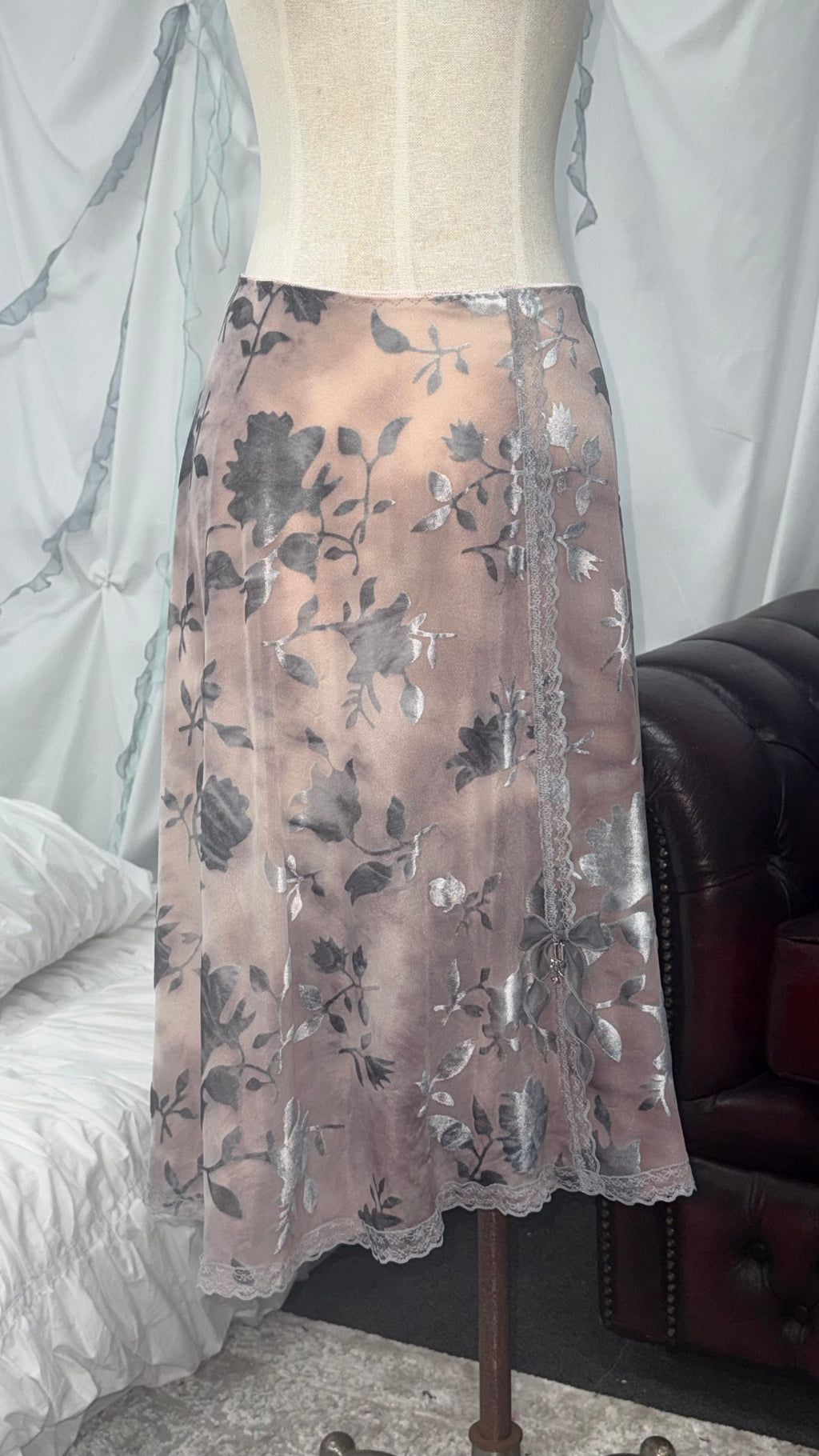 A sheer midi skirt with a gray floral pattern and scalloped lace trim, displayed on a cream-colored dress form in an indoor setting.