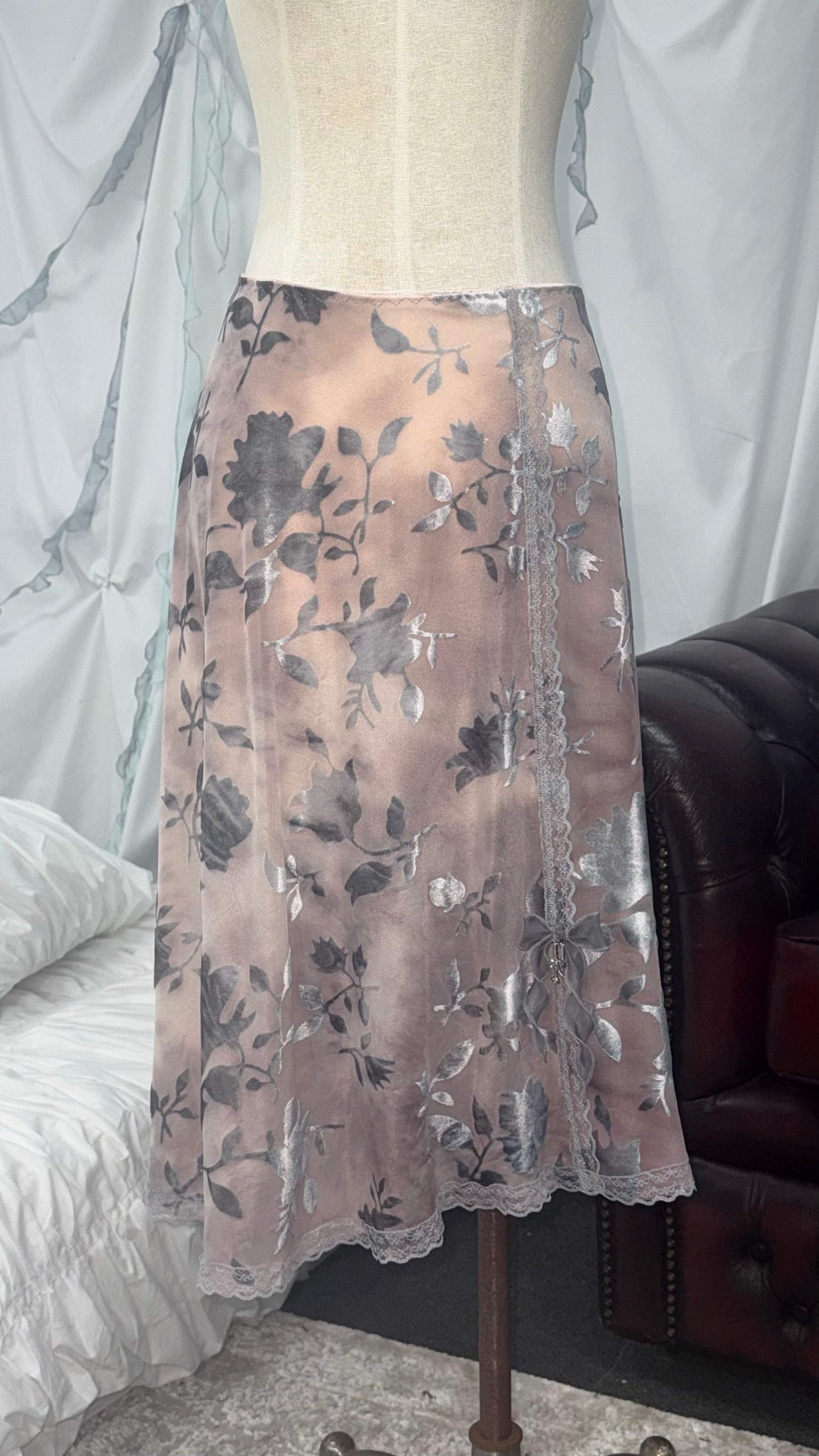A sheer midi skirt with a gray floral pattern and scalloped lace trim, displayed on a cream-colored dress form in an indoor setting.