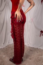 A model wearing a deep red lace maxi dress with a ruffled side overlay and tie details, standing against a draped white fabric backdrop.