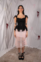 A model stands against a draped white fabric backdrop wearing a black ruffled sweetheart top with sheer cap sleeves and a light pink sheer midi skirt, paired with black lace-up platform shoes.