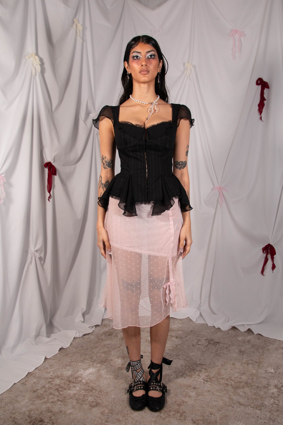 A model stands against a draped white fabric backdrop wearing a black ruffled sweetheart top with sheer cap sleeves and a light pink sheer midi skirt, paired with black lace-up platform shoes.