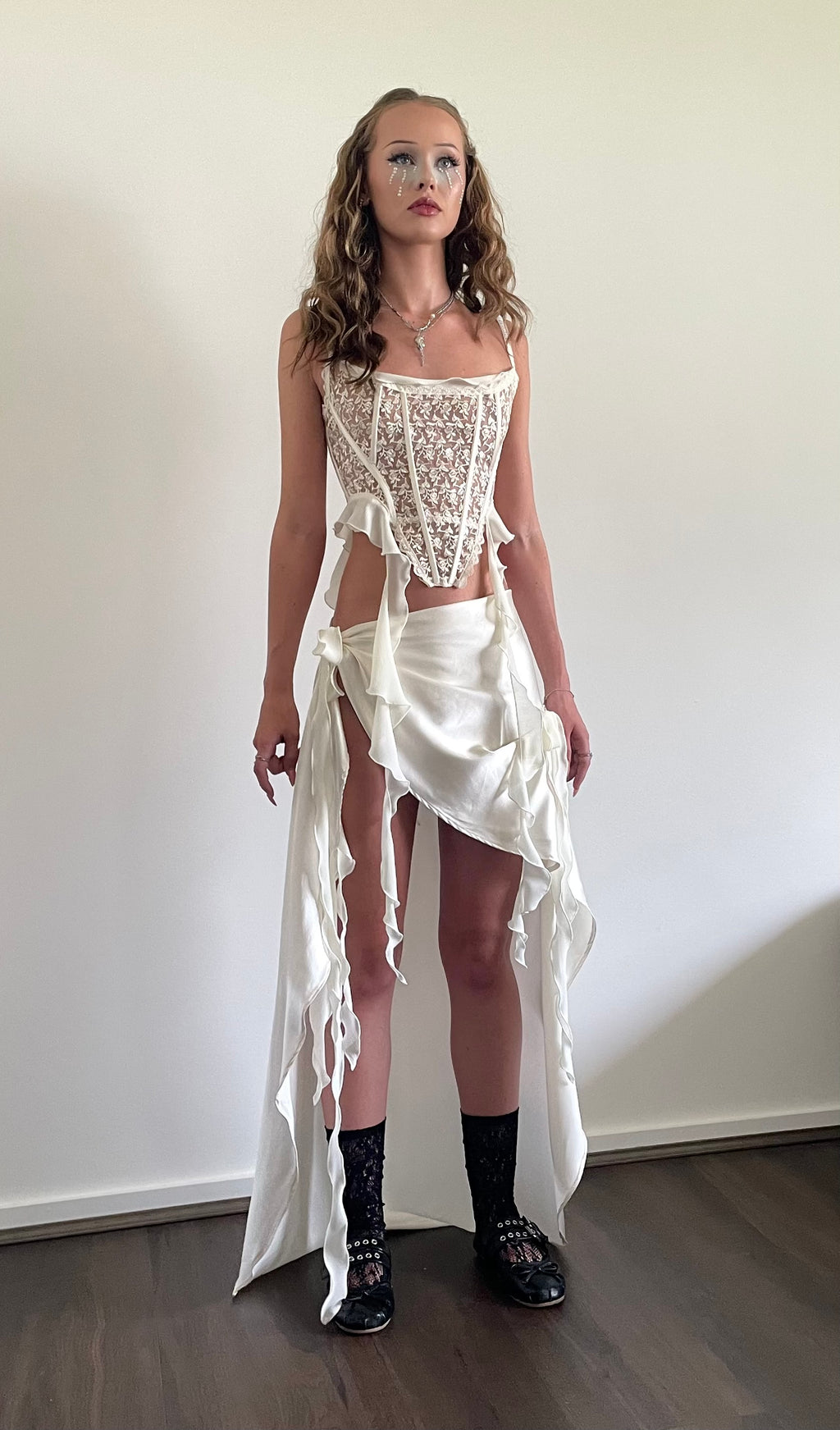A model stands in a room wearing a white lace corset top with a matching draped white fabric skirt or sarong, paired with black socks and black lace-up shoes.