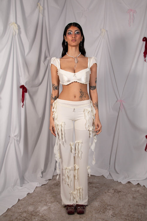 A model stands against a draped white fabric backdrop wearing a white two-piece outfit consisting of a ruffled crop top and matching distressed wide-leg pants with hanging tassel details.