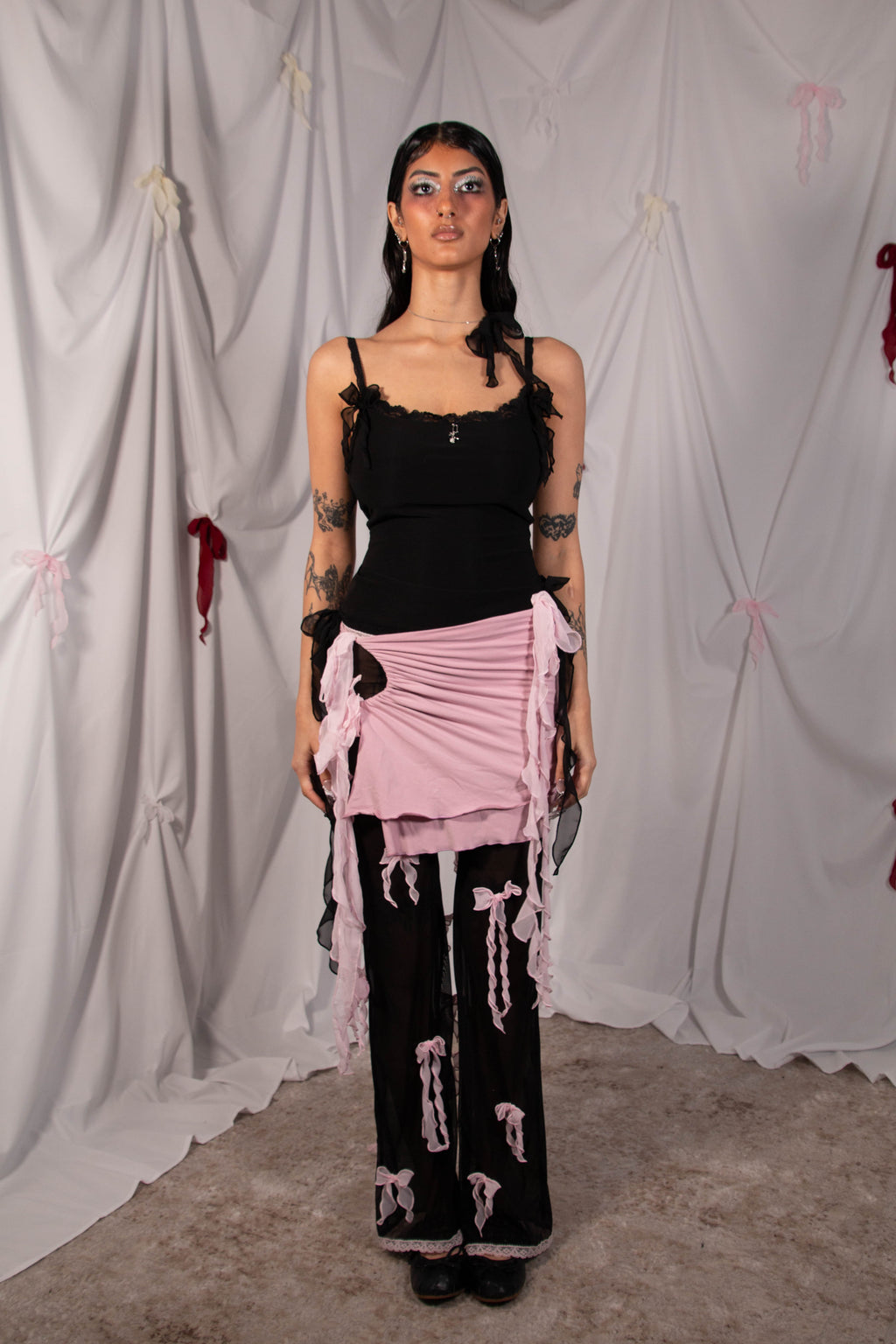 A model stands in front of a draped white fabric backdrop wearing a black strappy top, black wide-leg pants with pink jester motifs, and a pink ruffled sash wrapped around the waist.