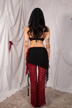 A woman shown from the back wearing a black head covering, black bralette, black wrap skirt with fringe, and red lace flare pants, standing in front of a white draped fabric backdrop.