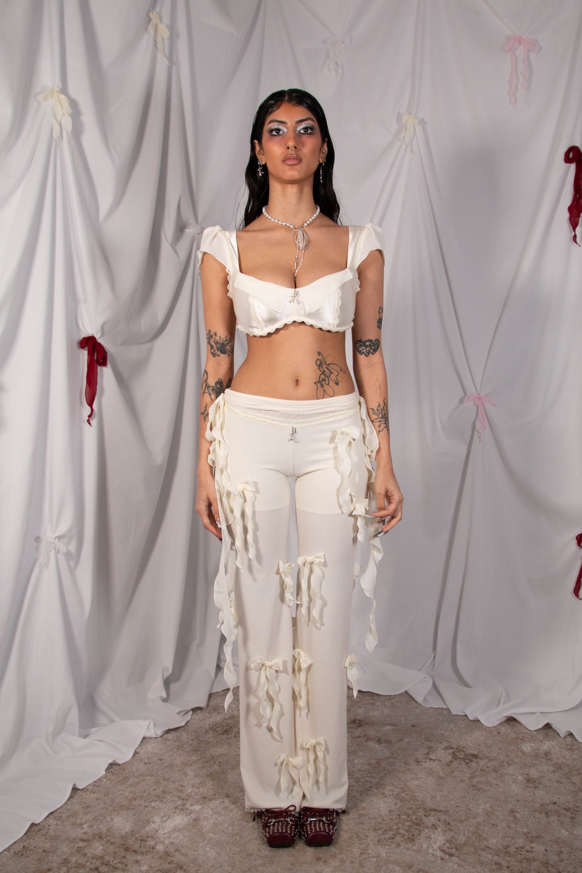 A model stands against a draped white fabric backdrop wearing a white two-piece outfit consisting of a ruffled crop top and matching distressed wide-leg pants with hanging tassel details.