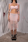 A model wearing a light pink two-piece outfit consisting of a sweetheart bra top with shoulder ties and a matching sheer polka-dot midi skirt, posed against a draped white fabric backdrop.