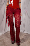 A person wearing sheer red lace flare pants with side ties and a red top, standing against a draped white fabric backdrop.