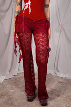 A person wearing sheer red lace flare pants with side ties and a red top, standing against a draped white fabric backdrop.