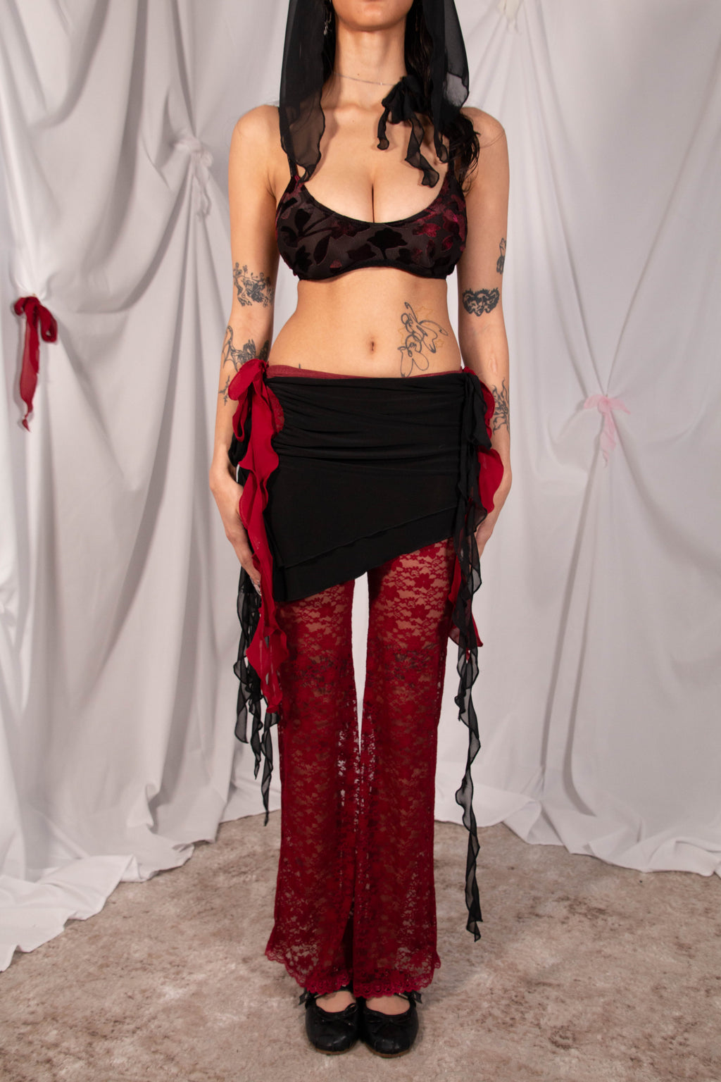 A model wearing a gothic-style two-piece outfit consisting of a black and red lace bralette top and matching red lace flared pants with black ruffled ties, standing against a white draped fabric backdrop.