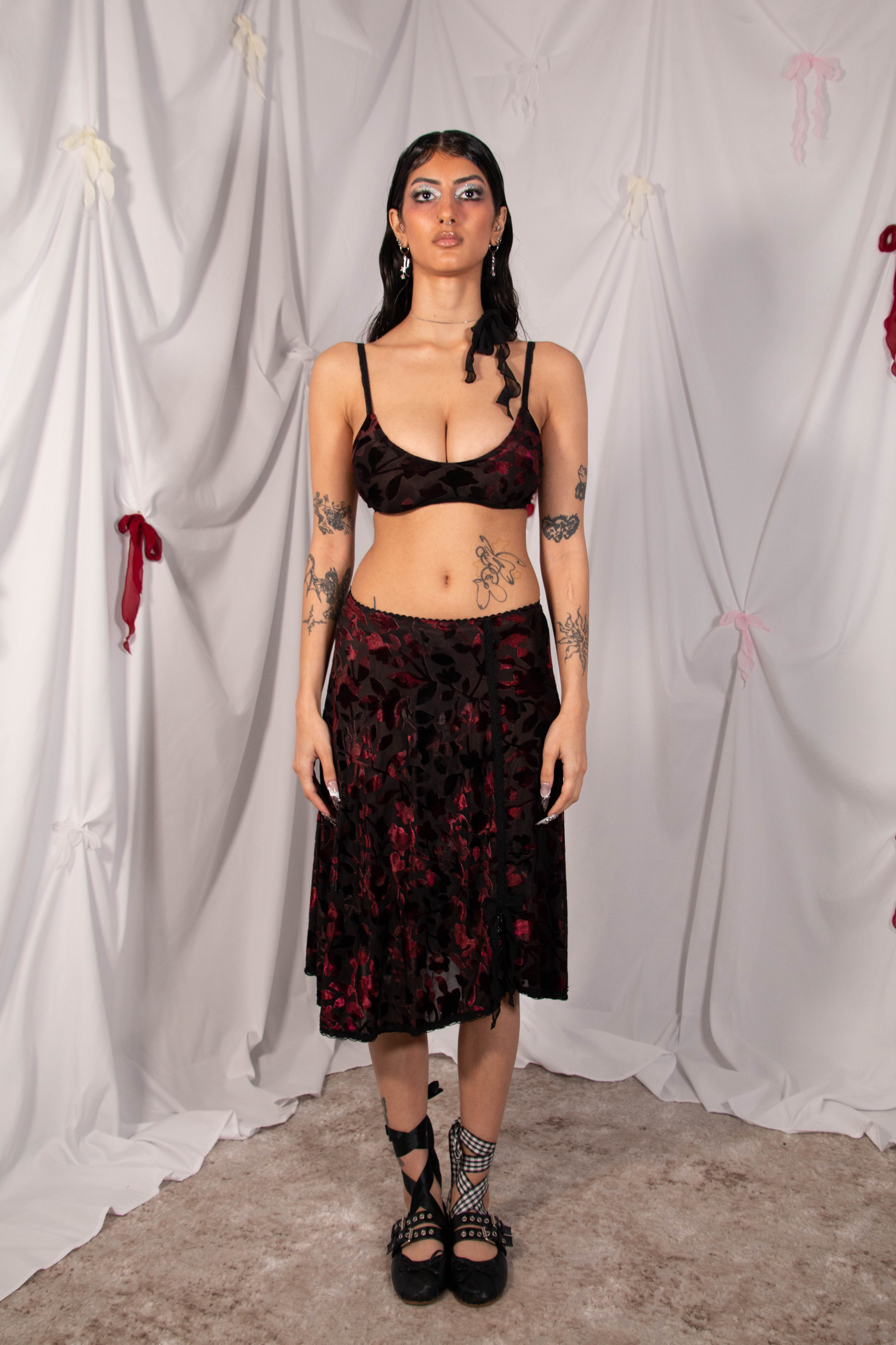 A model stands against a white draped fabric backdrop wearing a black and red floral print two-piece set consisting of a spaghetti-strap crop top and a matching midi skirt, paired with black lace-up platform shoes.