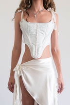 A woman modeling a white two-piece outfit consisting of a structured corset-style top with shoulder ties and a matching high-low wrap skirt with a side tie, paired with black lace-up shoes against a plain indoor wall.