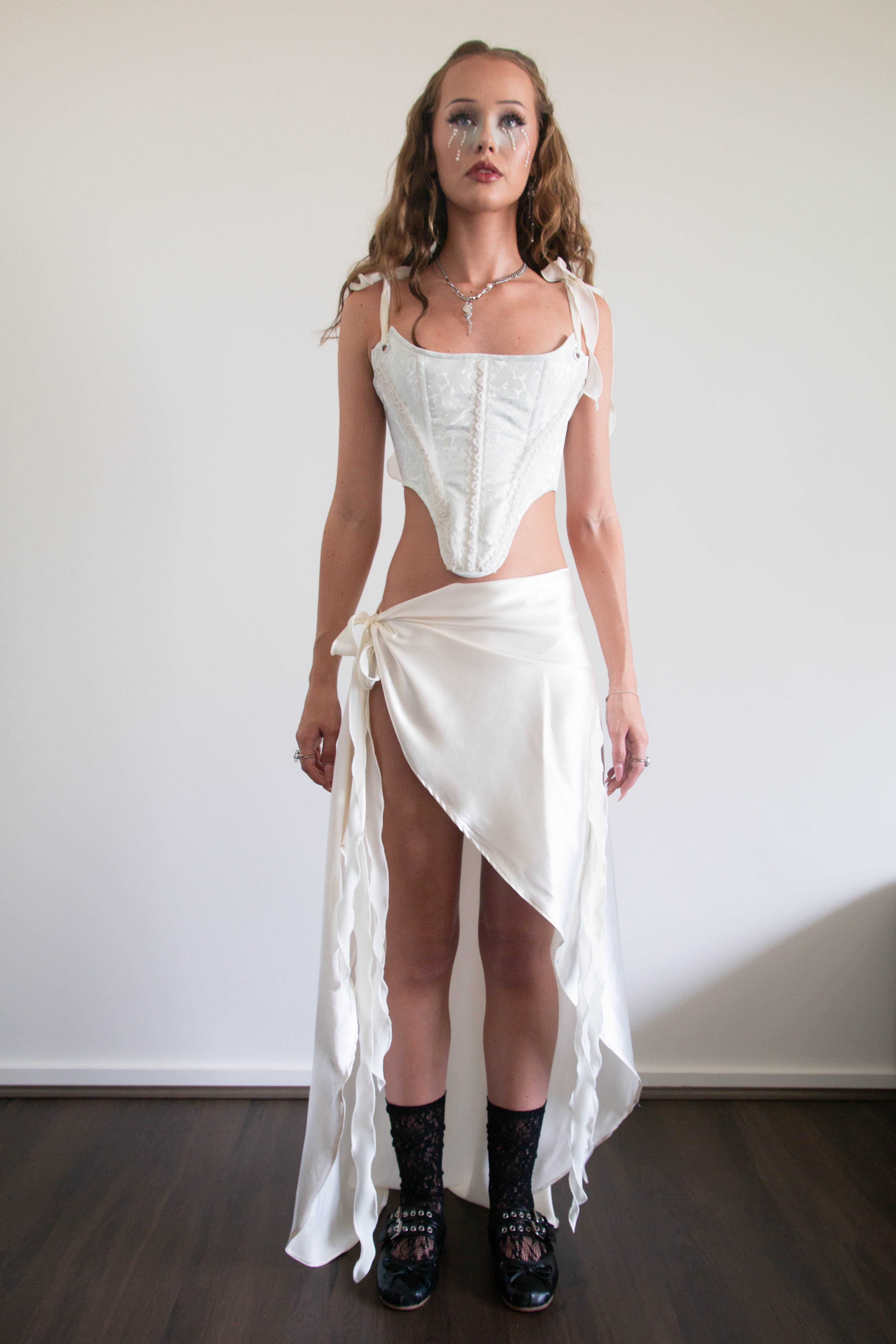 A woman modeling a white two-piece outfit consisting of a structured corset-style top with shoulder ties and a matching high-low wrap skirt with a side tie, paired with black lace-up shoes against a plain indoor wall.