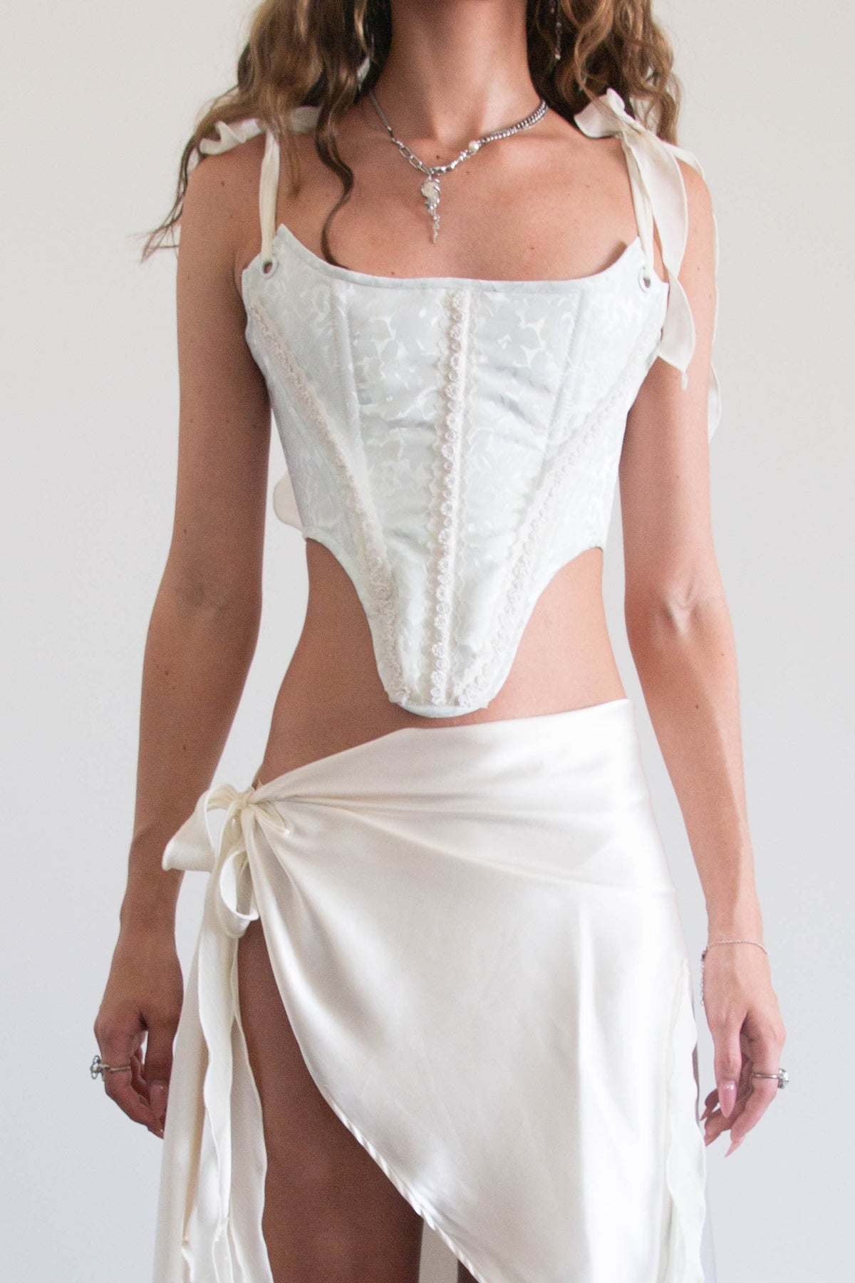 A woman modeling a white two-piece outfit consisting of a structured corset-style top with shoulder ties and a matching high-low wrap skirt with a side tie, paired with black lace-up shoes against a plain indoor wall.